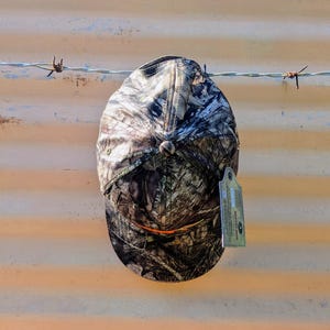 Op de afbeelding: Een camouflage baseballpet met een gebogen klep en een knoop aan de bovenkant. De pet heeft een patroon van groen, bruin en beige. Er is een label aan de pet bevestigd. De pet hangt aan een prikkeldraad.