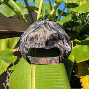 Op de afbeelding: Een camouflage baseballpet met een gebogen klep. De pet heeft een patroon van bruin, groen en bruin, ontworpen om op te gaan in buitenomgevingen. De achterkant van de pet heeft een verstelbare band voor een aangepaste pasvorm.