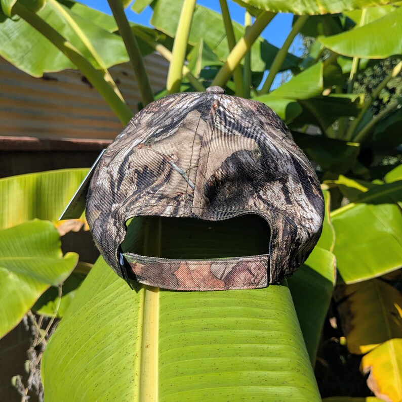 Op de afbeelding: Een camouflage baseballpet met een gebogen klep. De pet heeft een patroon van bruin, groen en bruin, ontworpen om op te gaan in buitenomgevingen. De achterkant van de pet heeft een verstelbare band voor een aangepaste pasvorm.