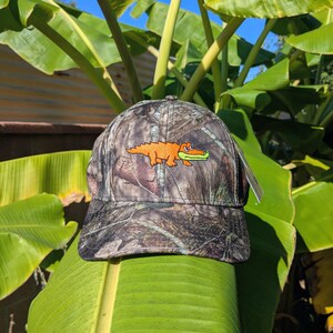Op de afbeelding: Camouflage baseballpet met een geborduurde oranje alligator en groene snuit. De pet heeft een gebogen klep en een label. De achtergrond toont groene bananenbladeren en een blauwe lucht.