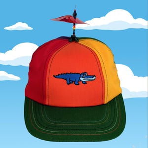 Könnte beinhalten: Bunte Baseballkappe mit Propeller. Die Kappe hat einen grünen Schirm und eine regenbogenfarbene Krone mit einem blauen Alligator, der auf der Vorderseite aufgestickt ist. Der Propeller ist rot und schwarz, und der Hintergrund ist ein blauer Himmel mit Wolken.