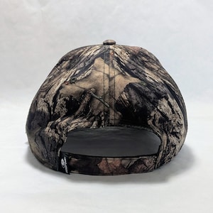 Op de afbeelding: Een camouflage baseballpet met een gebogen klep. De pet heeft een patroon van bruine, groene en bruine kleuren, dat lijkt op een bosrijke omgeving. De pet heeft een zwarte verstelbare band en een klein logo aan de zijkant.
