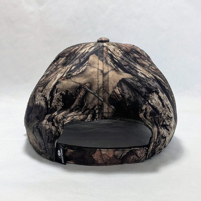 Op de afbeelding: Een camouflage baseballpet met een gebogen klep. De pet heeft een patroon van bruine, groene en bruine kleuren, dat lijkt op een bosrijke omgeving. De pet heeft een zwarte verstelbare band en een klein logo aan de zijkant.