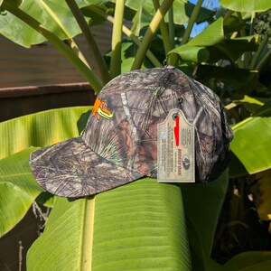 Op de afbeelding: Camouflage baseballpet met een bruin, groen en beige patroon. De pet heeft een oranje alligatorlogo en een hanglabel. Geschikt voor buitenactiviteiten.