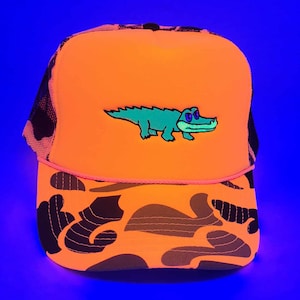 Könnte beinhalten: Leuchtend orangefarbene Trucker-Cap mit einem grünen Alligator-Design auf der Vorderseite. Die Kappe hat ein Camouflage-Muster auf dem Schirm und eine schwarze Mesh-Rückseite. Ein auffälliges Accessoire.