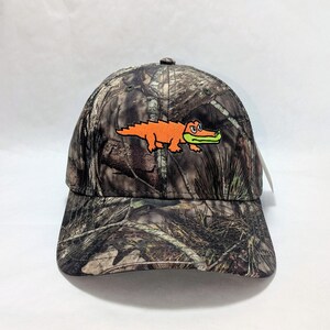 Op de afbeelding: Camouflage baseball cap met een oranje alligator ontwerp. De cap heeft een gebogen klep en een klassieke zes-panelen constructie. De alligator is geborduurd op de voorkant, wat een vleugje kleur toevoegt aan het bospatroon.