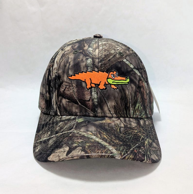Op de afbeelding: Camouflage baseball cap met een oranje alligator ontwerp. De cap heeft een gebogen klep en een klassieke zes-panelen constructie. De alligator is geborduurd op de voorkant, wat een vleugje kleur toevoegt aan het bospatroon.