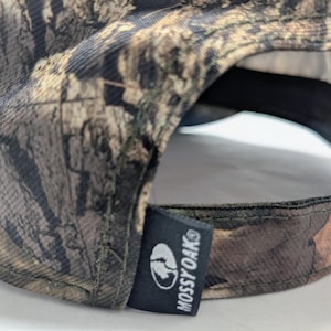 Op de afbeelding: Een camouflage baseballpet met een bruin, groen en beige patroon. De pet heeft een zwart label met het witte Mossy Oak logo en tekst.