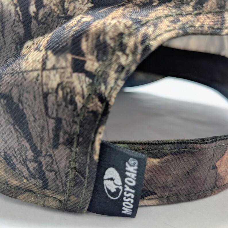 Op de afbeelding: Een camouflage baseballpet met een bruin, groen en beige patroon. De pet heeft een zwart label met het witte Mossy Oak logo en tekst.