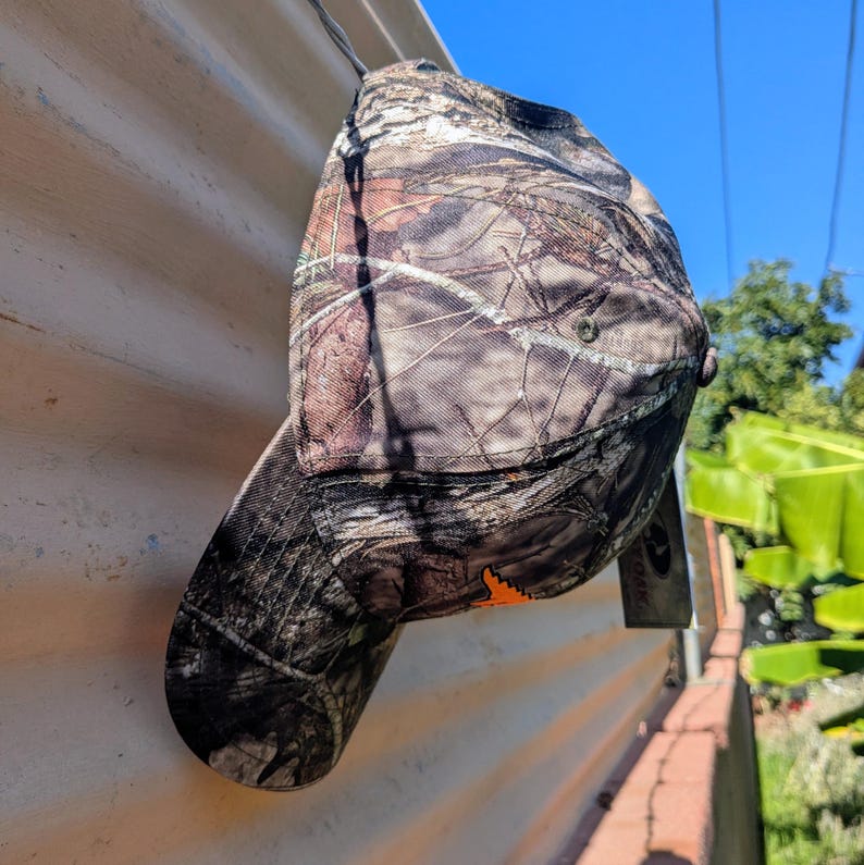 Op de afbeelding: Een camouflage baseballpet met een gebogen klep, in bruine, groene en beige tinten. De pet heeft een oranje logo en een label. Het hangt tegen een gegolfd metalen oppervlak. Geschikt voor buitenactiviteiten.