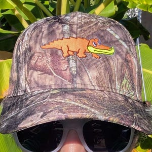 Op de afbeelding: Camouflage baseballpet met een geborduurde oranje alligator met groene bek. De pet heeft een gebogen klep en een bruin en groen camouflagepatroon. Een klassieke stijl, geschikt voor buitenactiviteiten.
