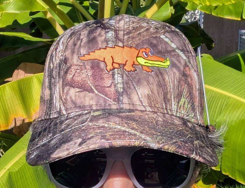 Op de afbeelding: Camouflage baseballpet met een geborduurde oranje alligator met groene bek. De pet heeft een gebogen klep en een bruin en groen camouflagepatroon. Een klassieke stijl, geschikt voor buitenactiviteiten.