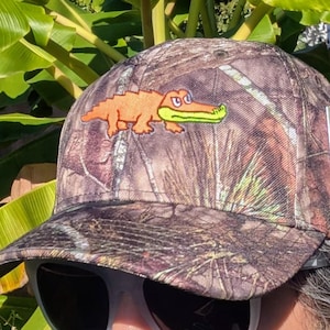 Op de afbeelding: Camouflage baseballpet met een oranje alligator patch en een groene bek. De pet heeft een gebogen klep en een Mossy Oak label. De hoed is ontworpen met een bos camouflage patroon.