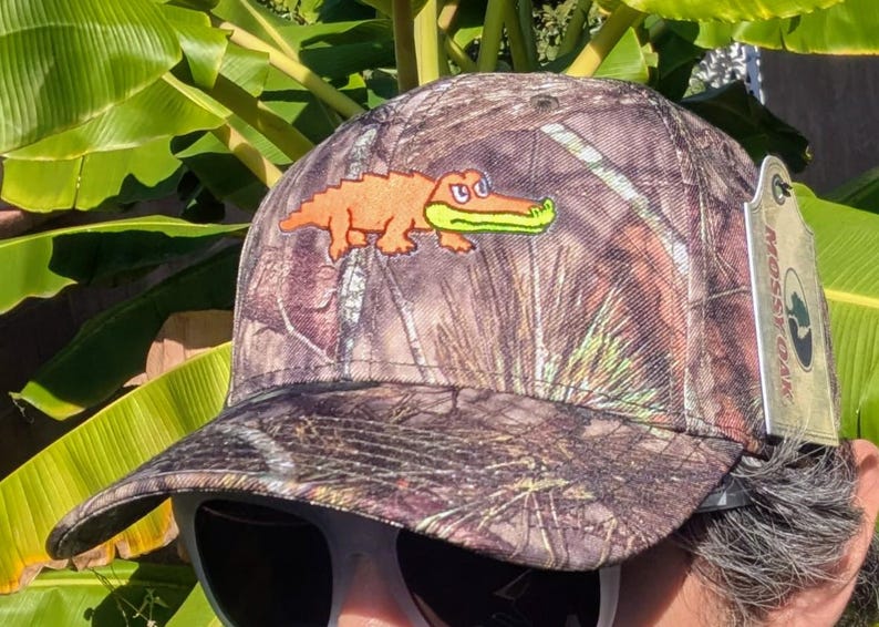 Op de afbeelding: Camouflage baseballpet met een oranje alligator patch en een groene bek. De pet heeft een gebogen klep en een Mossy Oak label. De hoed is ontworpen met een bos camouflage patroon.
