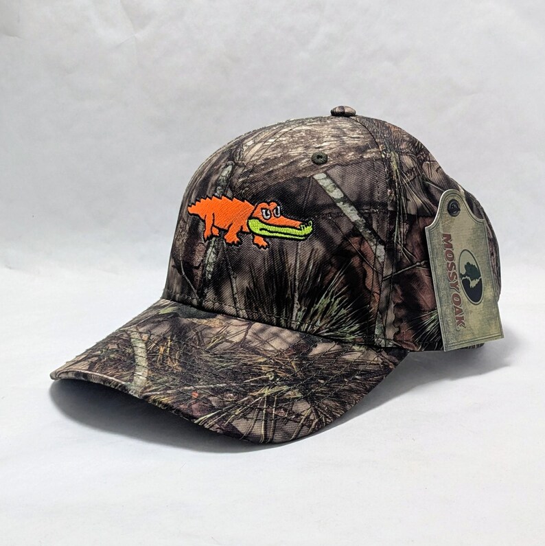 Op de afbeelding: Een camouflage baseball cap met een geborduurde oranje alligator en een groene bek. De cap heeft een gebogen klep en een label met de tekst "Mossy Oak".