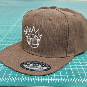 May include: A brown and white baseball cap with a white graphic of a cartoon character with a crown and a large mouth. The cap has a black and white label that reads "The Pioneer of Authentic Headwear Snapback High Quality Flat Premium Headwear".