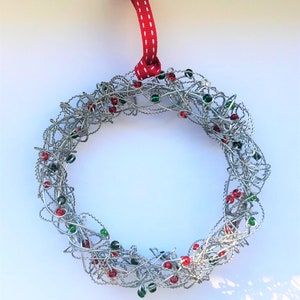 Op de afbeelding: Een zilveren draadkrans met rode en groene kralen, versierd met een rood lint met witte stippen.