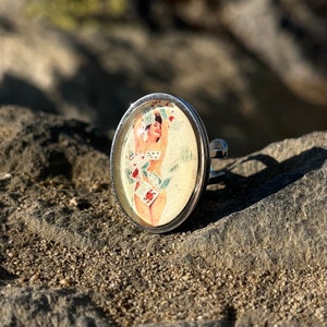 May include: A silver ring with a glass cabochon featuring a vintage illustration of a woman in a swimsuit. The woman is surrounded by playing cards and flowers.