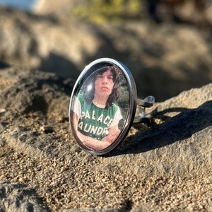 May include: A silver ring with a round, clear glass cabochon featuring a photo of a person wearing a green shirt with the text "Palace Launder" printed on it.