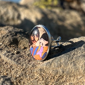 Könnte beinhalten: Ein silberner Ring mit einem runden, klaren Glascabochon, der ein Vintage-Foto von zwei Männern zeigt. Die Männer tragen bunte Kleidung und haben langes Haar. Der Ring liegt auf einer felsigen Oberfläche.