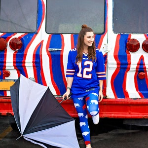 May include: A woman wearing a blue jersey with the number "12" and white and red buffalo print leggings stands next to a red, white, and blue striped bus. She is holding a black and white umbrella.