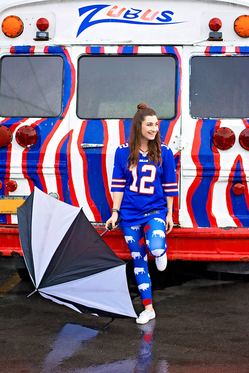 May include: A woman wearing a blue jersey with the number "12" and white and red buffalo print leggings stands next to a red, white, and blue striped bus. She is holding a black and white umbrella.