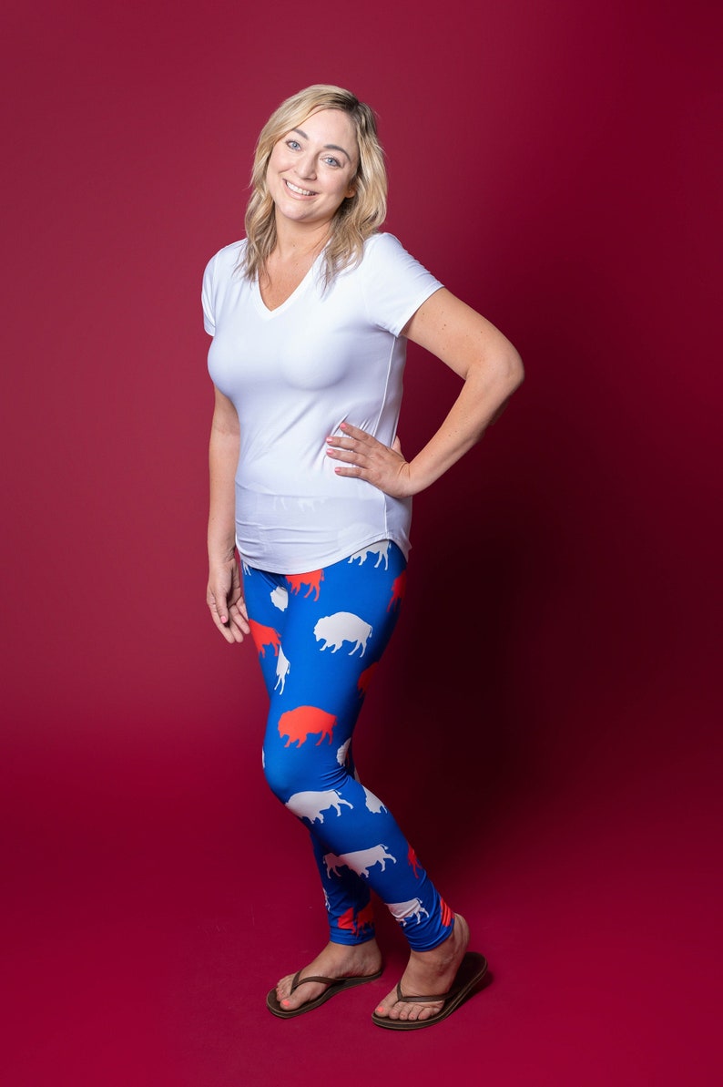 May include: A woman wearing a white v-neck t-shirt and blue leggings with a white and red buffalo print. She is standing on a red background.
