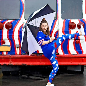 May include: A woman wearing blue and white leggings with a white and red animal print, a blue jersey, and white sneakers, stands in front of a red, white, and blue striped bus with the word "ZUBUS" written on the side. She is holding a black umbrella over her head.