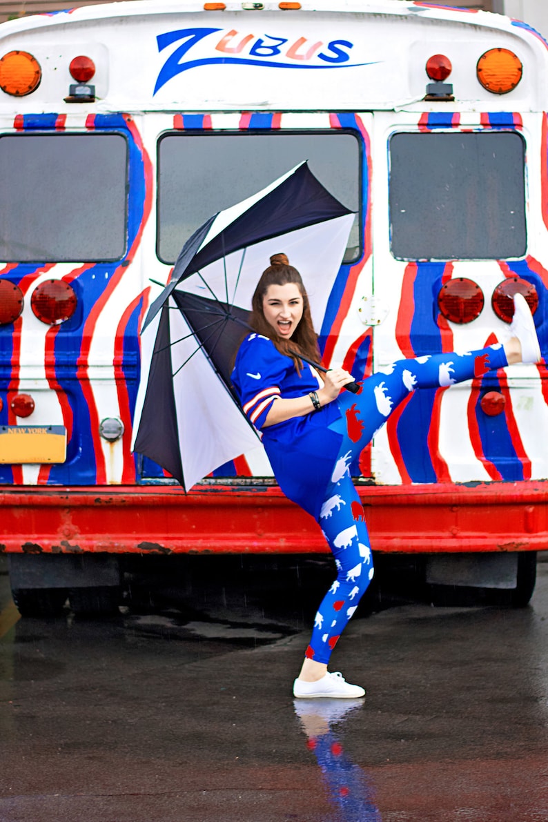 May include: A woman wearing blue and white leggings with a white and red animal print, a blue jersey, and white sneakers, stands in front of a red, white, and blue striped bus with the word "ZUBUS" written on the side. She is holding a black umbrella over her head.