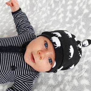 May include: A baby wearing a black and white striped onesie and a black knotted hat with a white buffalo print.