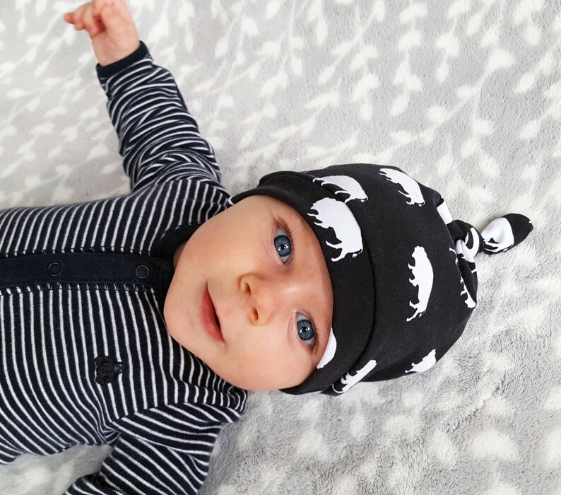 May include: A baby wearing a black and white striped onesie and a black knotted hat with a white buffalo print.