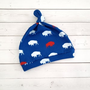 May include: Blue baby hat with a white buffalo pattern and a red buffalo in the center. The hat has a knot on top.
