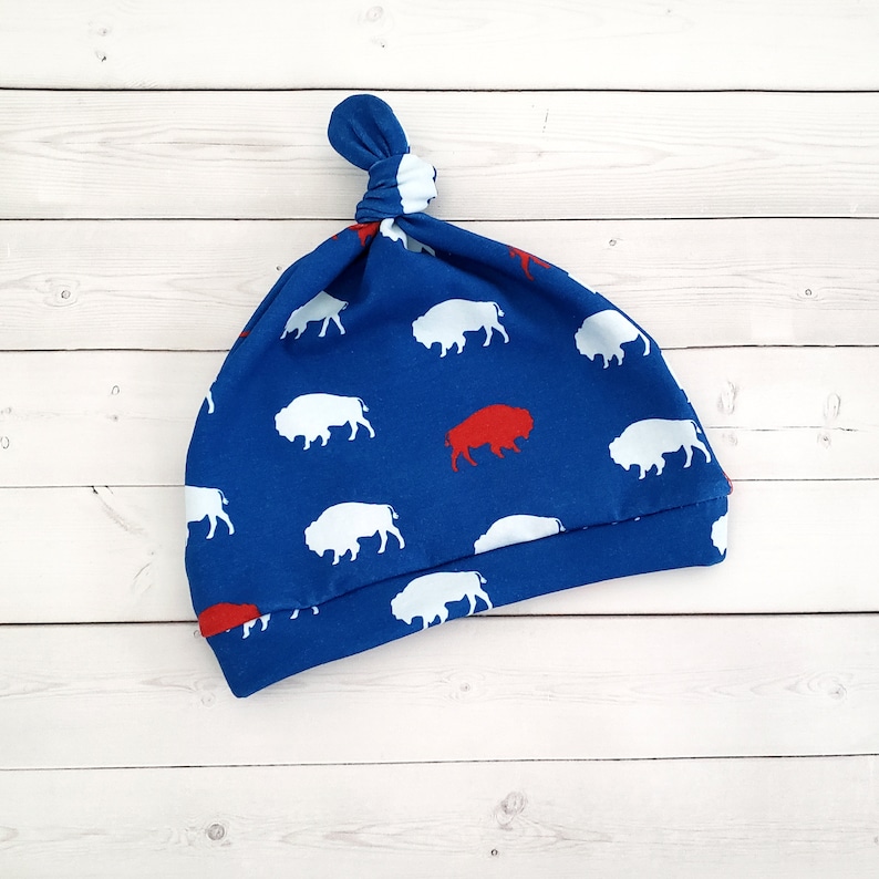 May include: Blue baby hat with a white buffalo pattern and a red buffalo in the center. The hat has a knot on top.