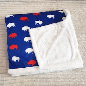 May include: A blue blanket with a pattern of red and white bison. The blanket has a soft, white, fluffy underside and a white border. The bison are evenly spaced across the blue fabric.