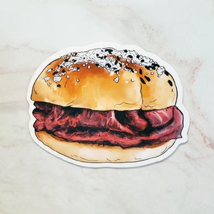 May include: A sticker of a cartoon illustration of a pastrami on rye sandwich with sesame seeds on the bun.
