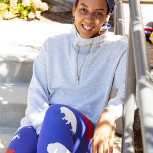 May include: A woman wearing a grey sweatshirt and blue leggings with a white buffalo print. She is sitting on a set of concrete steps with a railing behind her.