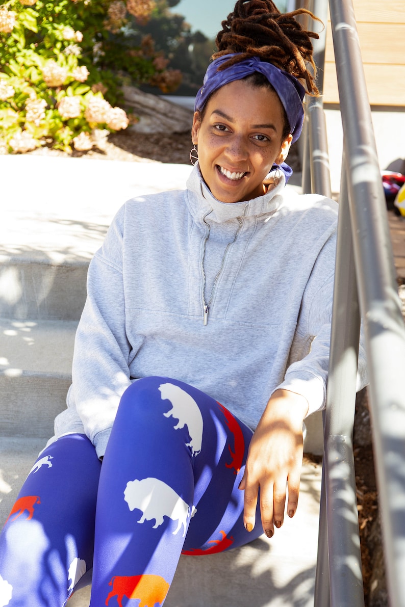 May include: A woman wearing a grey sweatshirt and blue leggings with a white buffalo print. She is sitting on a set of concrete steps with a railing behind her.