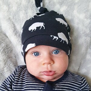 May include: A baby wearing a navy blue and white knotted hat with a buffalo print. The baby has blue eyes and is looking directly at the camera.