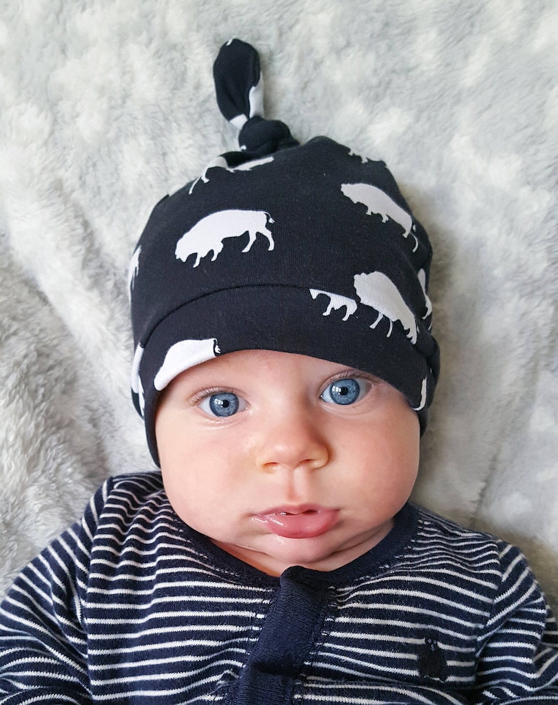 May include: A baby wearing a navy blue and white knotted hat with a buffalo print. The baby has blue eyes and is looking directly at the camera.