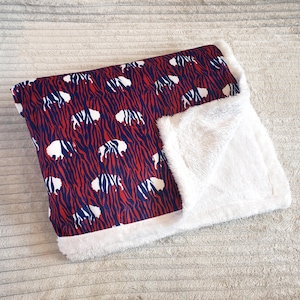 May include: A red, white, and blue patterned blanket featuring a buffalo design. The blanket has a soft, white, plush lining. The buffalo pattern is set against a red background with blue stripes.