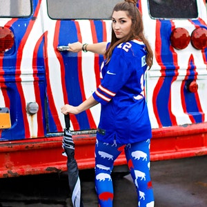May include: A woman wearing a blue jersey with white trim and white and red buffalo leggings stands in front of a red, white, and blue striped bus. She is holding a black and white umbrella.