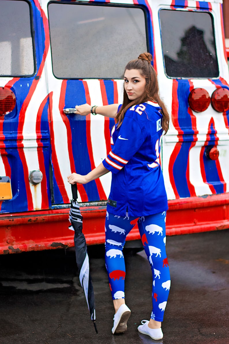 May include: A woman wearing a blue jersey with white trim and white and red buffalo leggings stands in front of a red, white, and blue striped bus. She is holding a black and white umbrella.