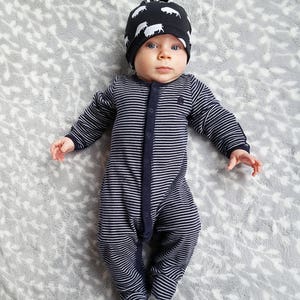 May include: A baby wearing a black and white striped sleeper and a black and white patterned hat with a knot on top. The baby is lying on a gray and white patterned blanket.