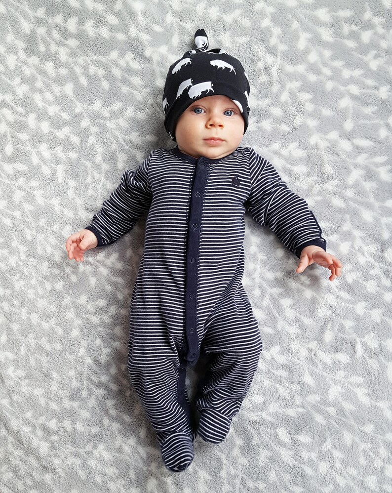 May include: A baby wearing a black and white striped sleeper and a black and white patterned hat with a knot on top. The baby is lying on a gray and white patterned blanket.