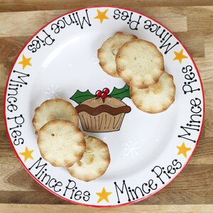 May include: A white ceramic plate with a red rim features a festive Christmas design. The plate is decorated with snowflakes, holly, and the words "Mince Pies". It has a collection of six small, round, golden-brown mince pies on top.