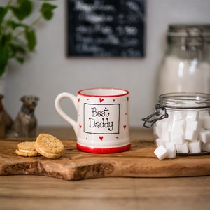 May include: White ceramic mug with red trim and red hearts. The mug has the text "Best Daddy" in black lettering. The mug is on a wooden cutting board with two cookies.