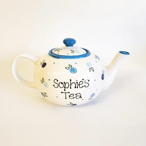 Könnte beinhalten: Weiße Keramik-Teekanne mit blauem Deckel und Griff. Die Teekanne ist mit blauen floralen Designs und dem Schriftzug "Sophie's Tea" verziert. Ausguss und Rand sind ebenfalls blau. Ein charmantes Objekt für Teeliebhaber.