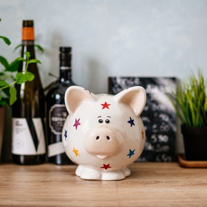Puede incluir: Huchita de cerámica blanca con estrellas de colores y un hocico rosa. La huchita está sobre una mesa de madera.