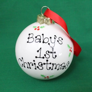 May include: White Christmas bauble with red ribbon and the text "Baby's 1st Christmas" in black lettering with holly leaf and berry accents.