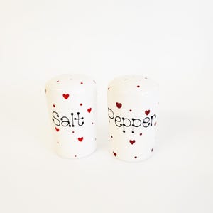 Puede incluir: Salero y pimentero de cerámica blanca con diseños de corazones rojos y letras negras. El salero dice "Salt" y el pimentero dice "Pepper".